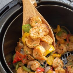 Wooden spoon lifting cooked shrimp and mixed vegetables from an air fryer basket.