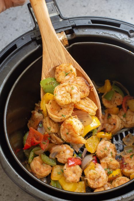 Wooden spoon lifting cooked shrimp and mixed vegetables from an air fryer basket.