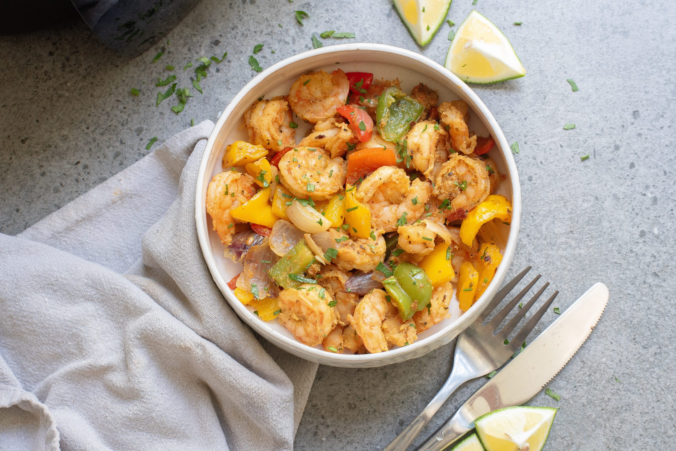 A bowl of shrimp stir-fry with bell peppers and onions, garnished with herbs. A cloth napkin, fork, spoon, and lime wedges are nearby on a gray surface.