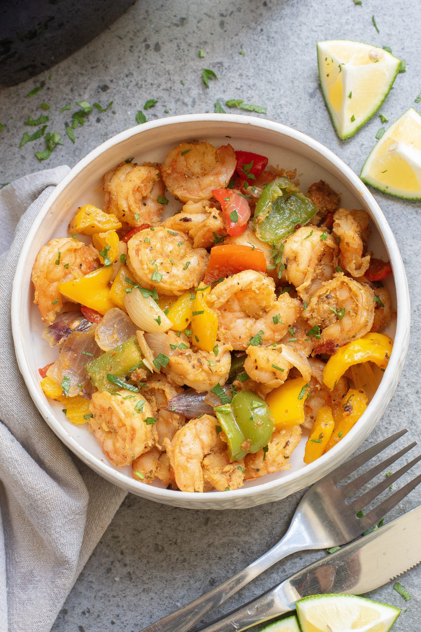 A bowl of Keto Fajita Shrimp Dinner with bell peppers and onions, garnished with herbs. Fork, knife, and lime wedges on the side.