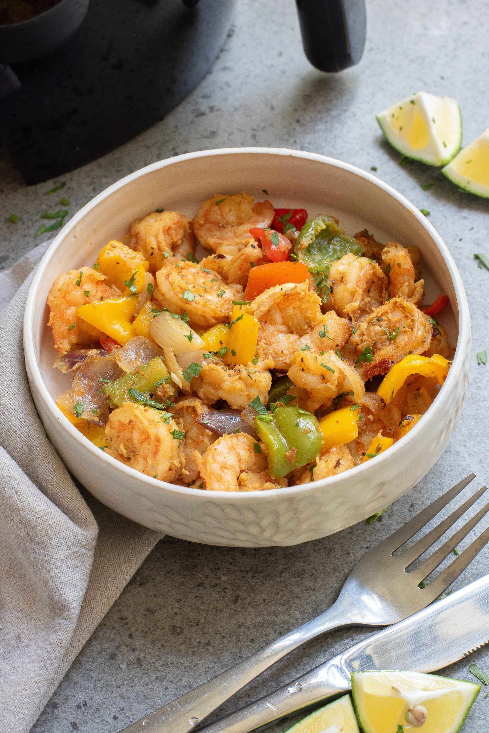 A bowl of Keto Fajita Shrimp Dinner with yellow and green bell peppers, onions, and herbs. Lime wedges, a fork, and a knife are nearby on a textured surface.