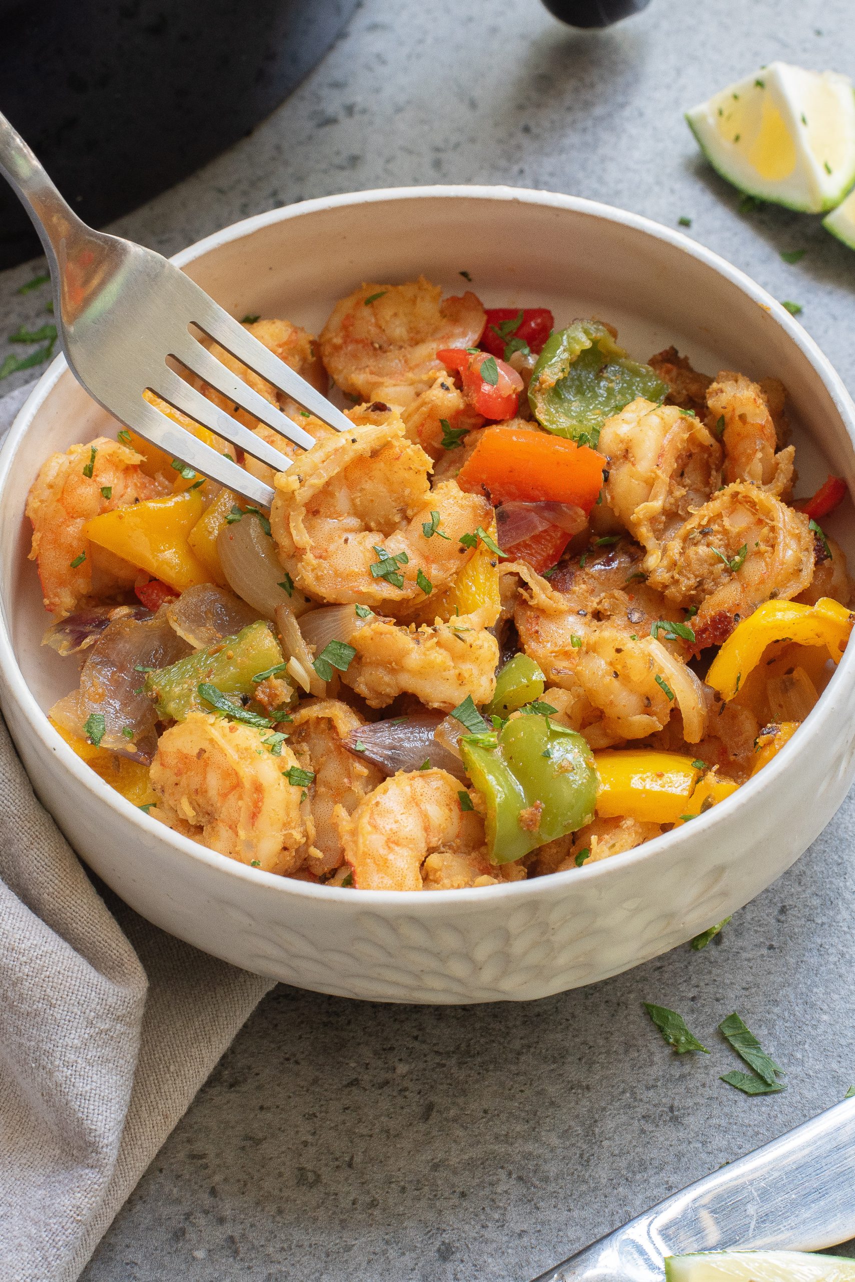 A fork in a bowl of cooked shrimp with colorful bell peppers and onions. Lime wedges are visible in the background.