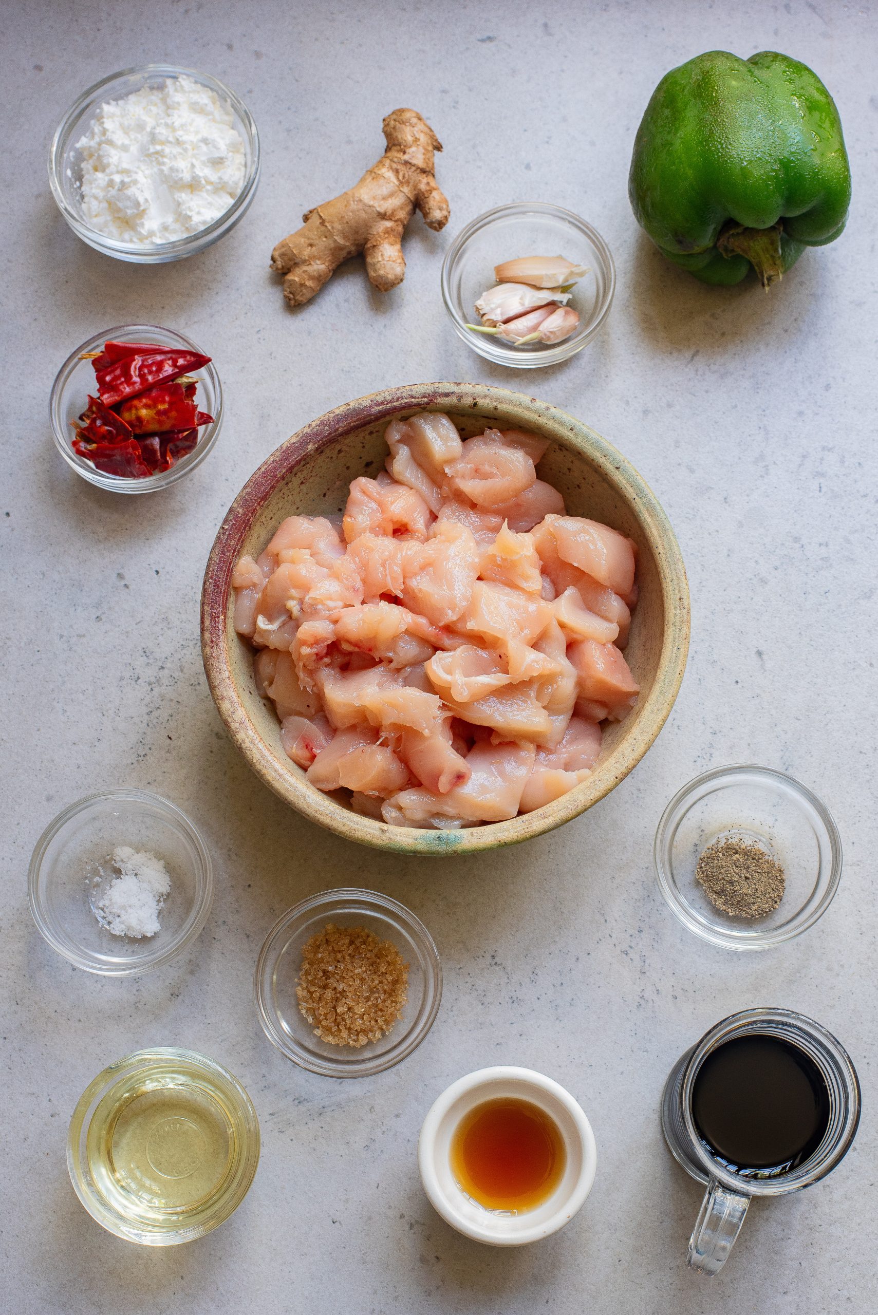 Ingredients for cooking: a bowl of diced chicken, ginger, green bell pepper, seasoning, and sauces arranged on a light surface.