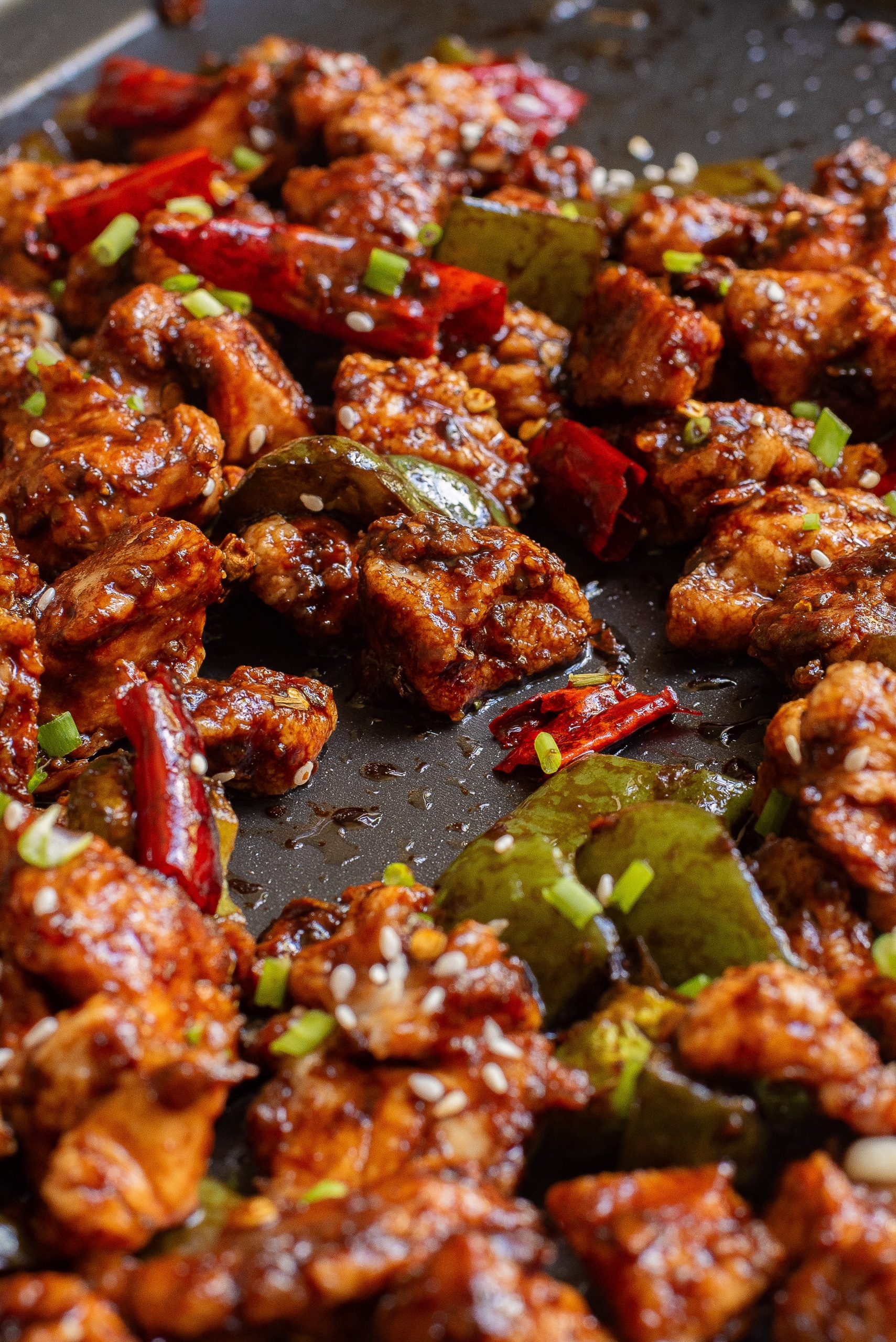 Close-up of a spicy stir-fry dish with pieces of meat, red chilies, and green peppers, garnished with sesame seeds and chopped green onions.