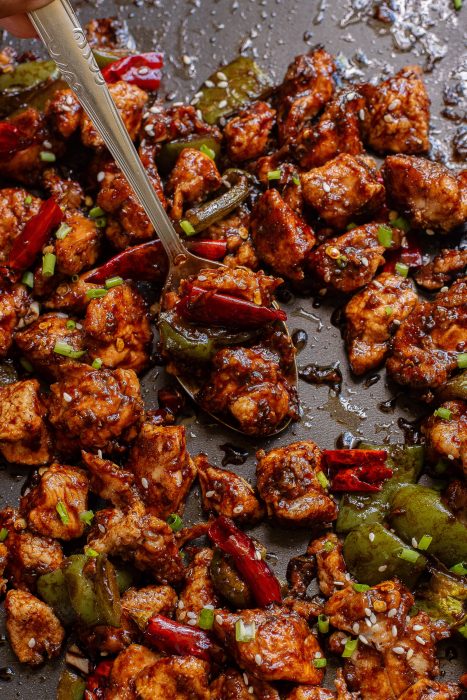 A pan filled with Baked Thai Garlic Chicken, diced, cooked chicken, green bell peppers, and red chilies, garnished with chopped green onions and sesame seeds, with a spoon placed on the dish.