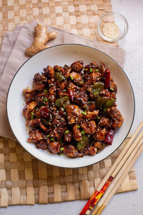 A bowl of fried chicken pieces with vegetables and spices, garnished with sesame seeds, placed on a woven mat alongside chopsticks and a small dish of sesame seeds.