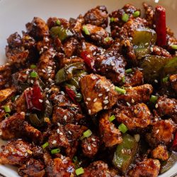 A bowl of stir-fried chicken with green peppers, red chilies, and a glazed sauce, garnished with sesame seeds and chopped green onions.