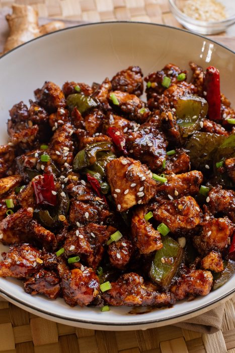 A bowl of stir-fried chicken with green peppers, red chilies, and a glazed sauce, garnished with sesame seeds and chopped green onions.