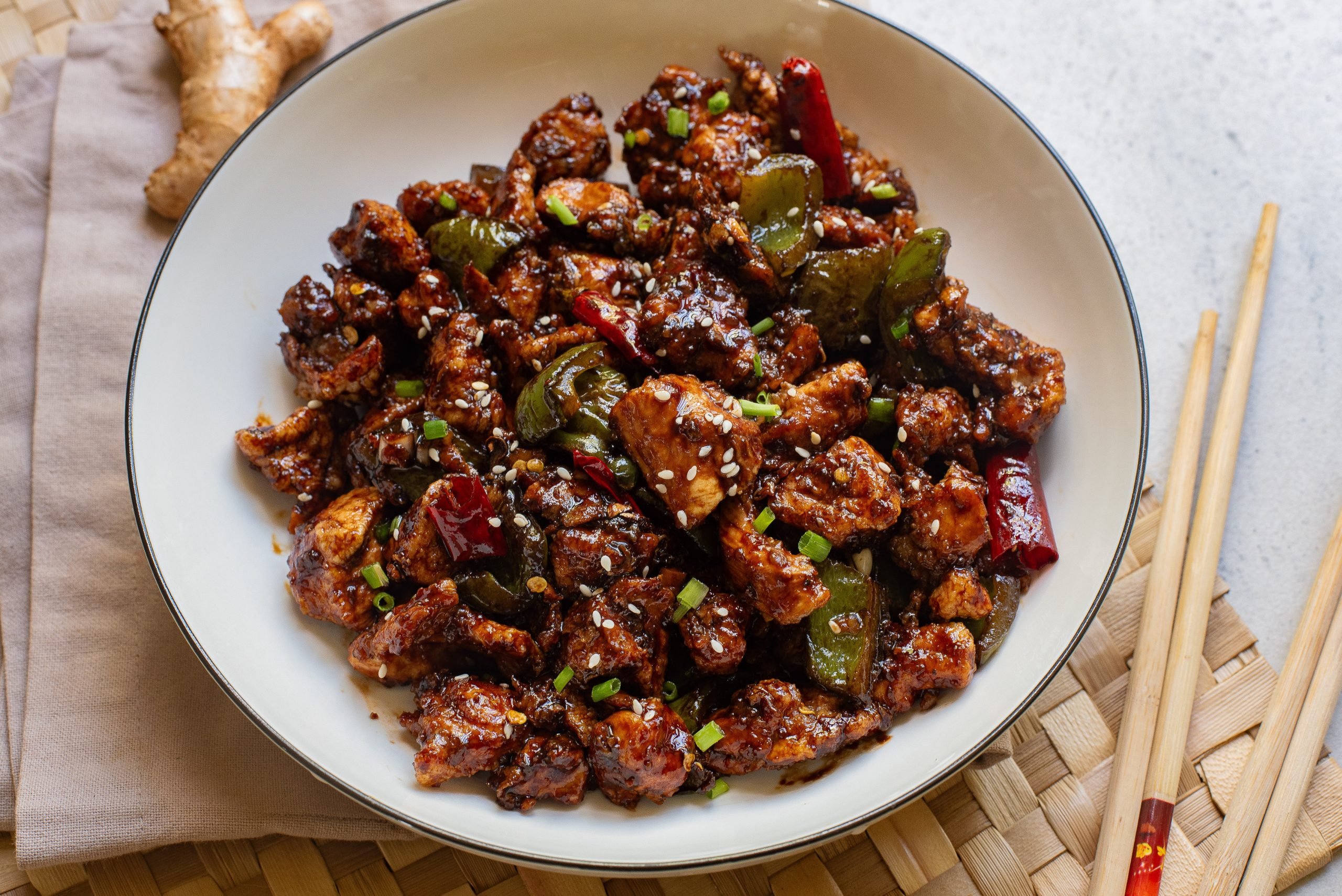 A bowl of spicy stir-fried chicken with green peppers and dried red chilies, garnished with sesame seeds, on a woven mat next to chopsticks and ginger.