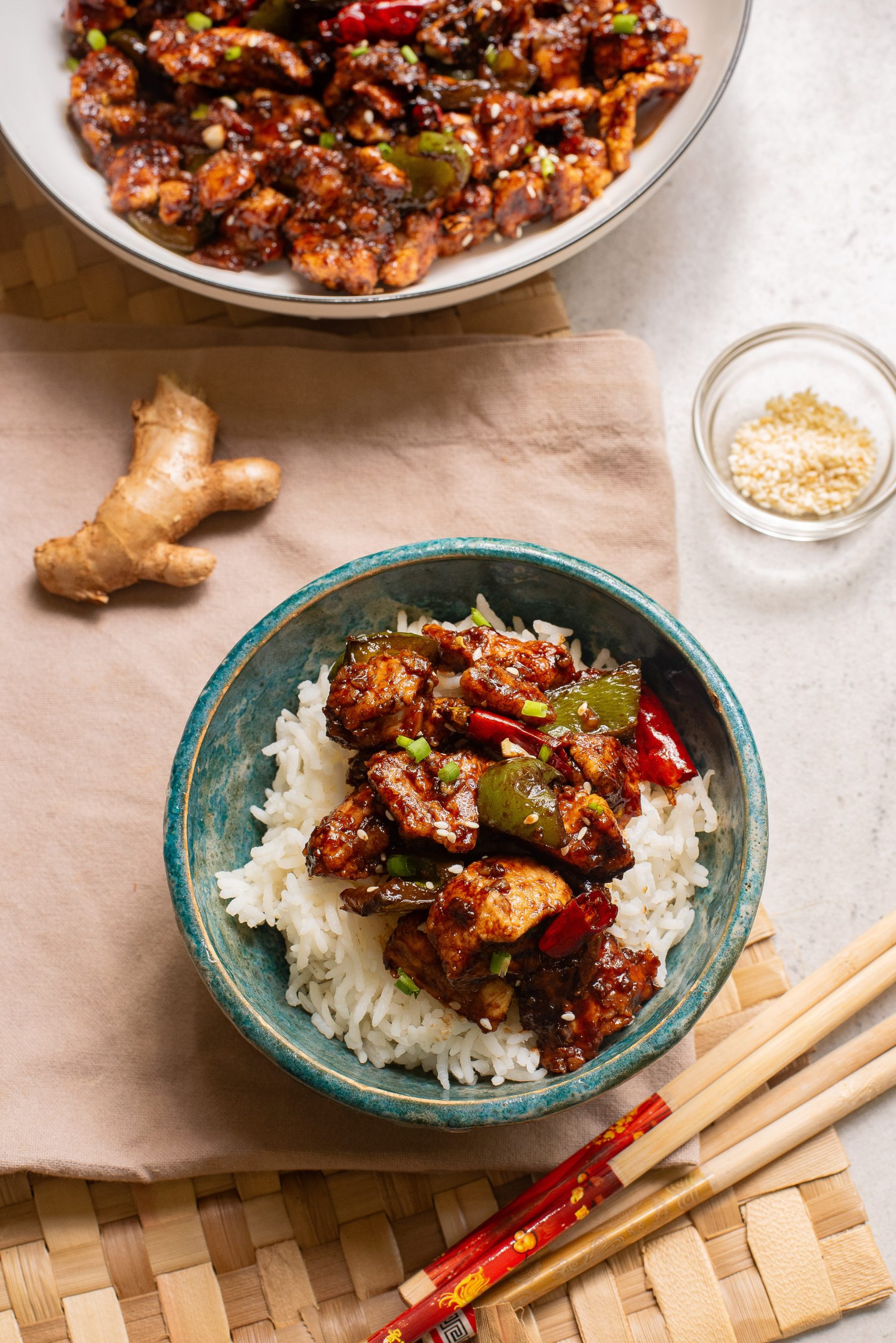 A blue bowl of Baked Thai Garlic Chicken topped with stir-fried chicken, bell peppers, and red chilies. Nearby are chopsticks, ginger, sesame seeds, and a larger bowl of the same dish.