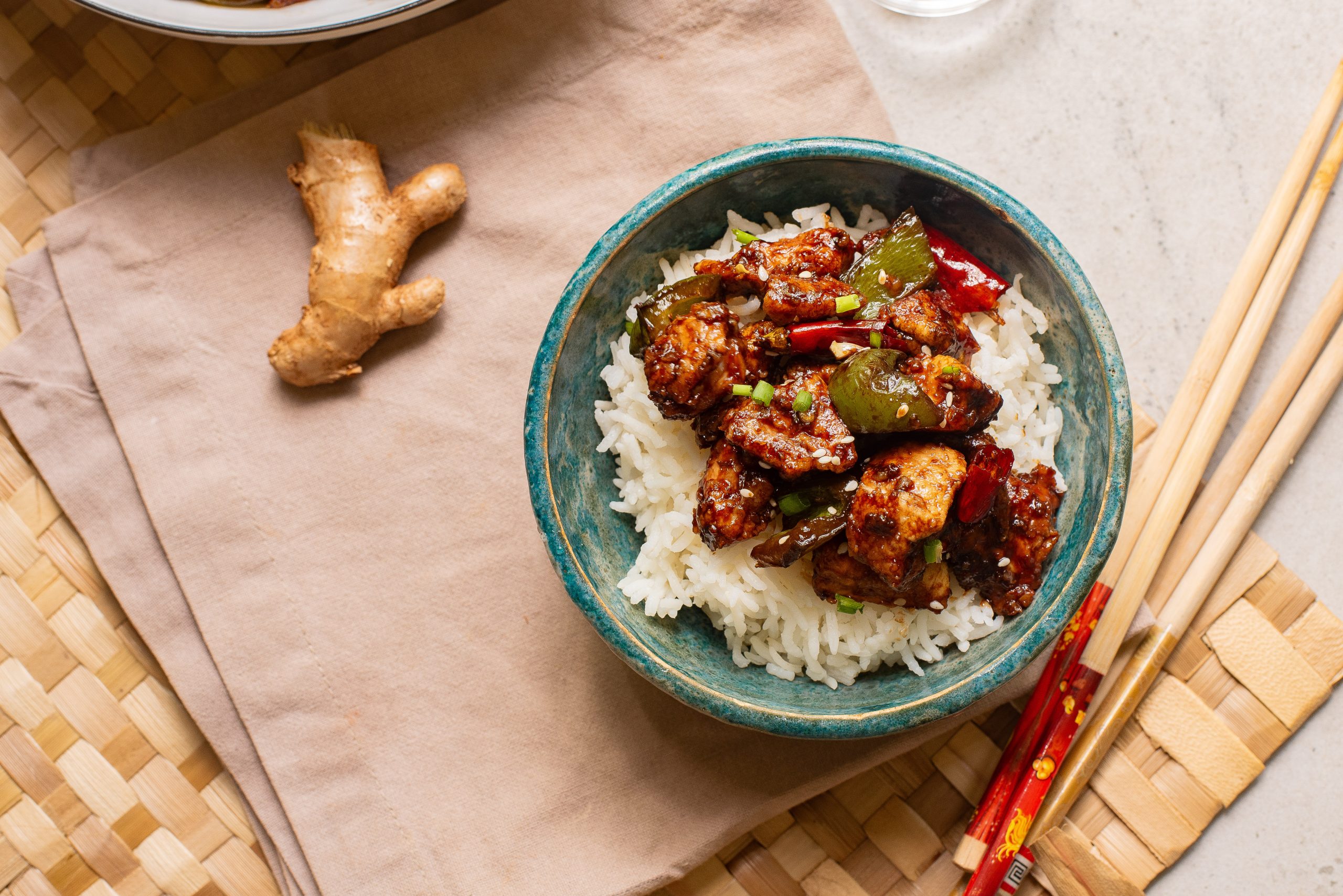 A bowl of rice topped with stir-fried chicken and vegetables, with ginger root and chopsticks nearby on a woven mat.