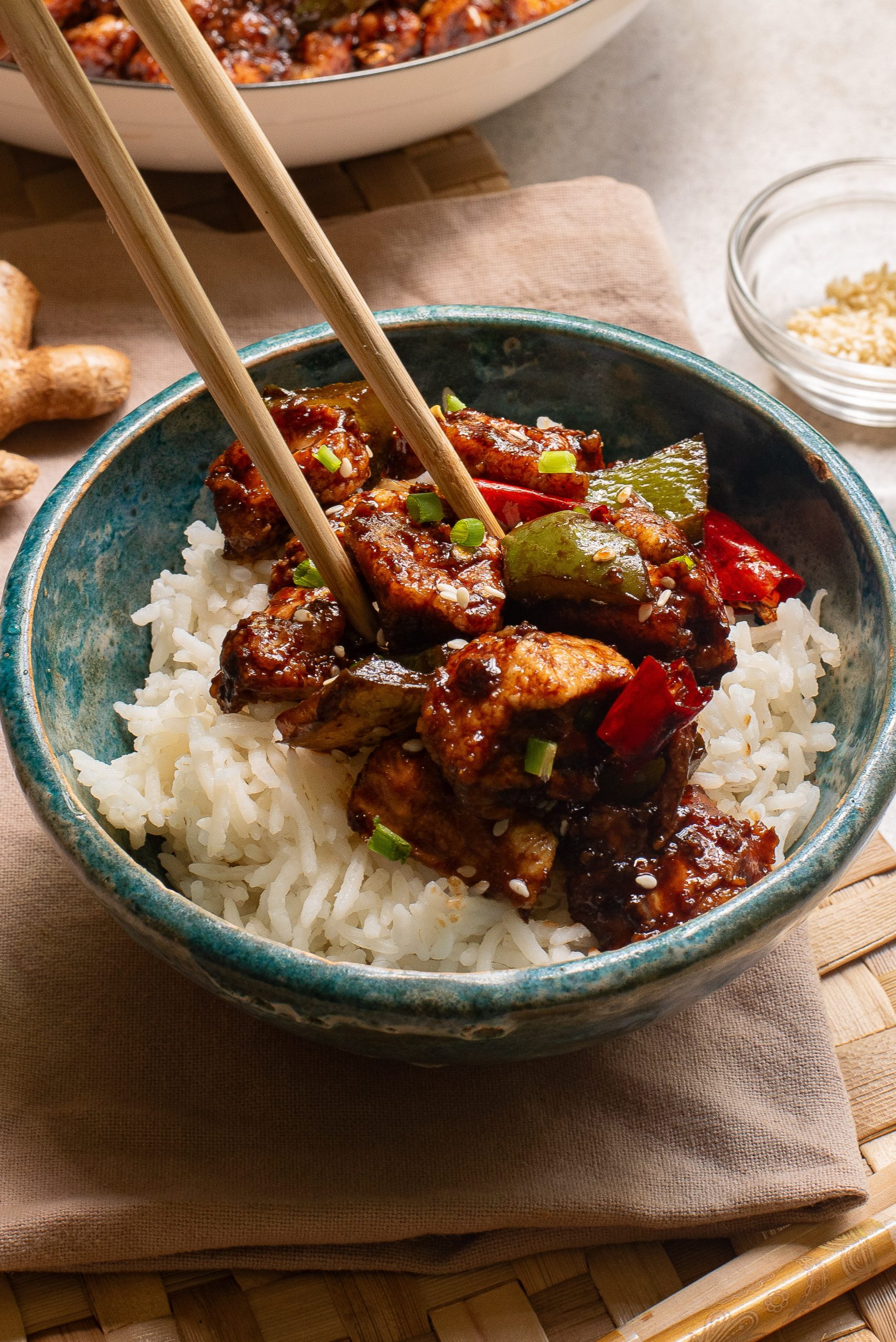 A blue bowl of Baked Thai Garlic Chicken topped with stir-fried vegetables and tofu, garnished with sesame seeds. Chopsticks rest in the bowl.