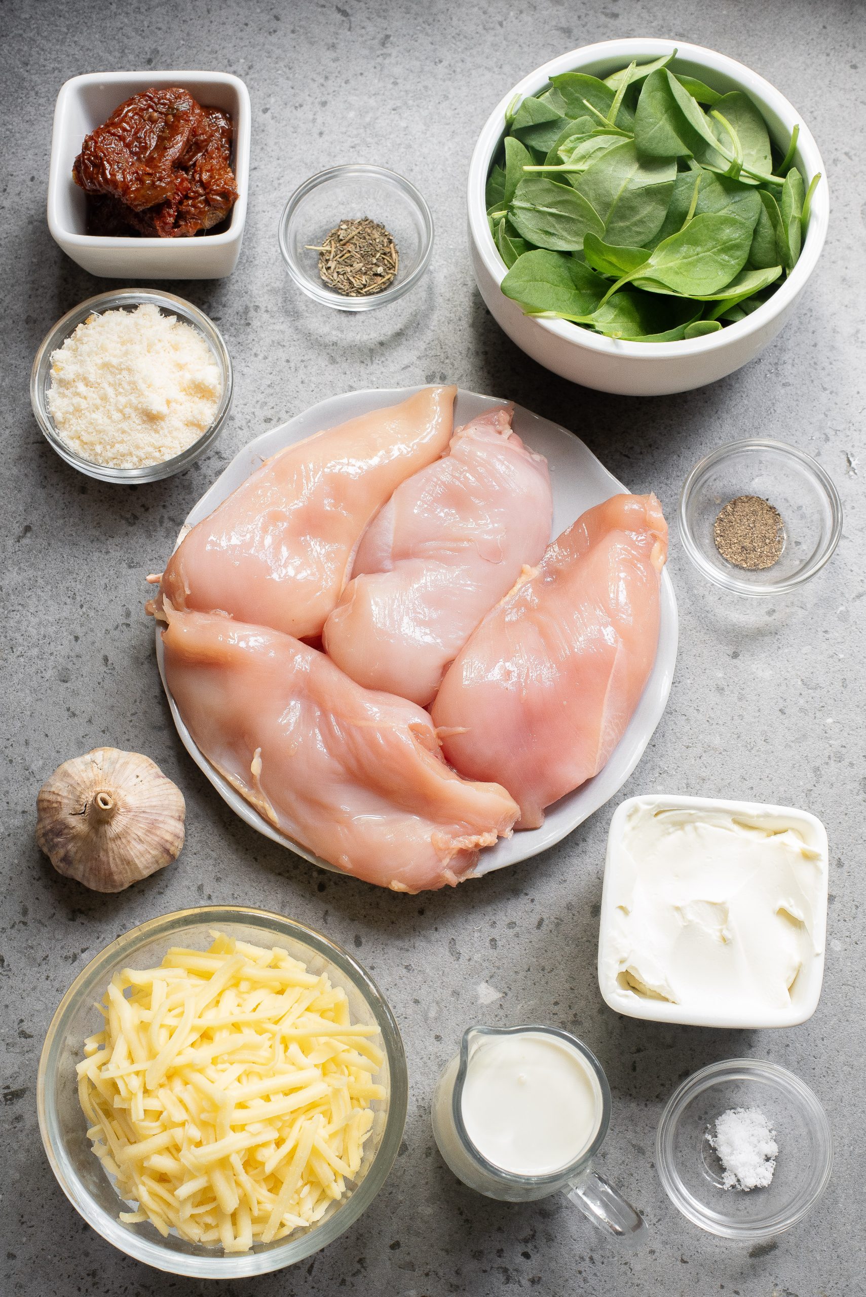 Ingredients on a plate surrounded by bowls of spinach, cream cheese, grated cheese, cream, garlic, sun-dried tomatoes, parmesan, and seasonings on a gray surface.