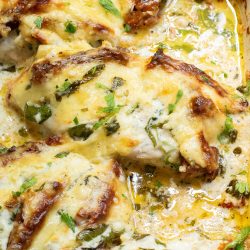 Baked chicken breasts covered in melted cheese and herbs, garnished with fresh parsley in a baking dish.