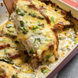 Baked dish with cheesy, herb-topped chicken breast being lifted from a red casserole dish on a gray surface.