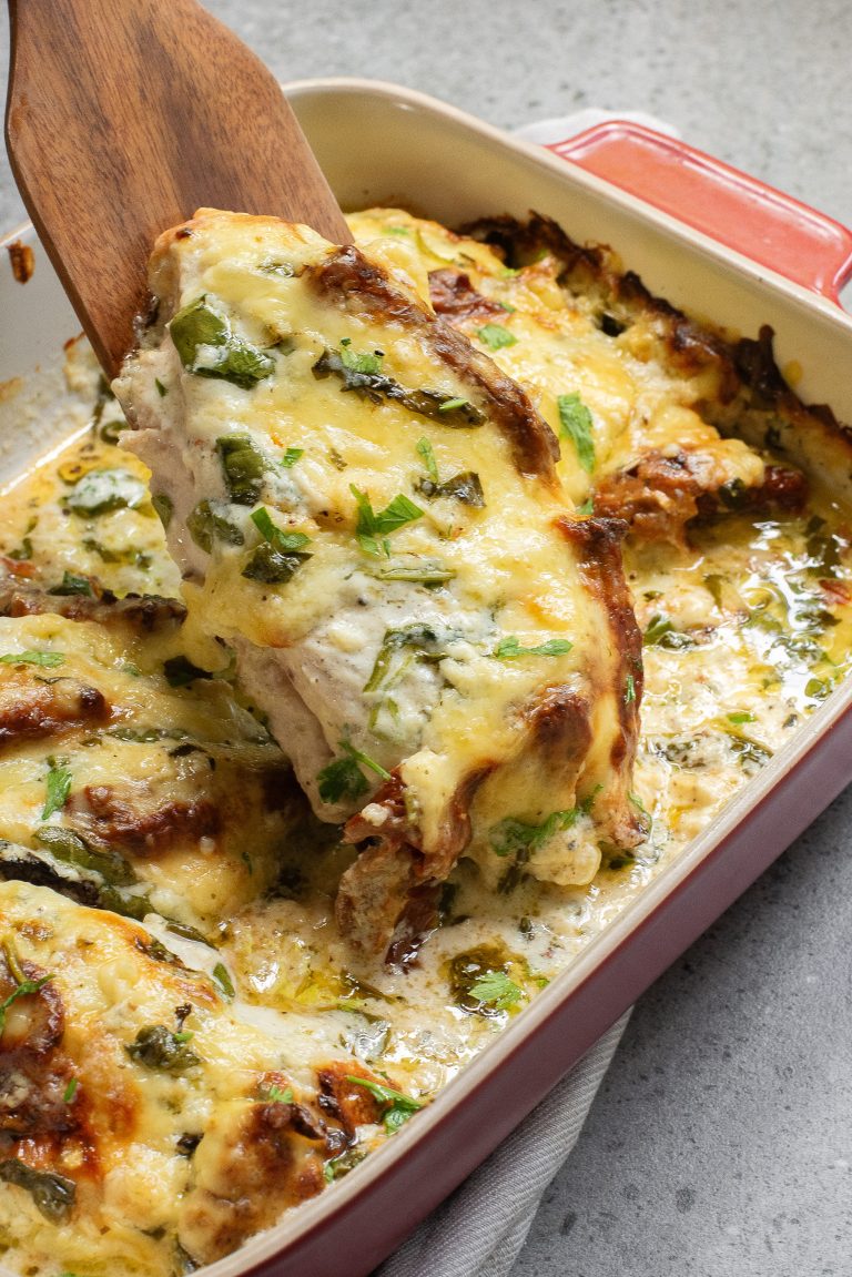 Baked dish with cheesy, herb-topped chicken breast being lifted from a red casserole dish on a gray surface.