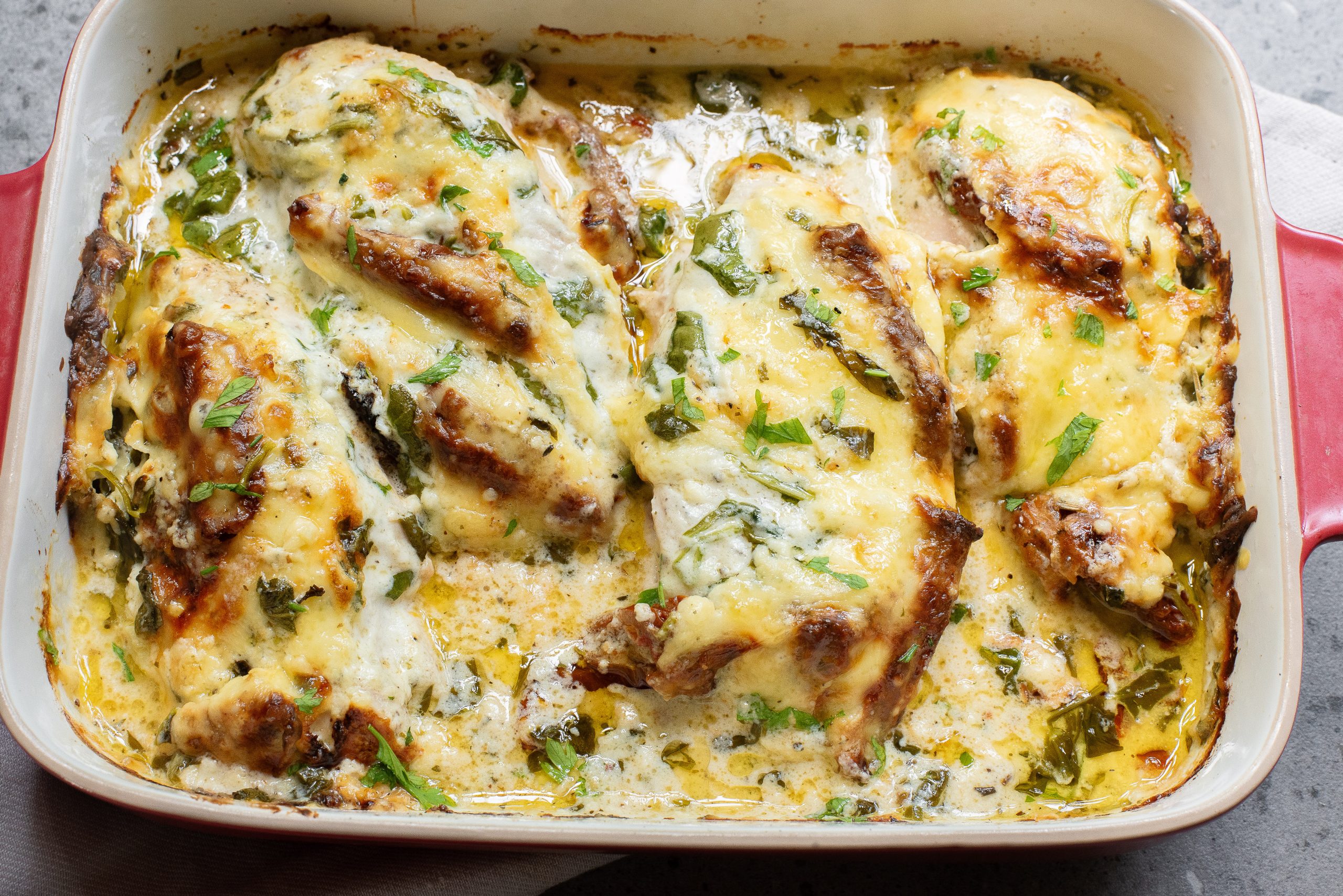 Baked chicken breasts in a red dish, topped with melted cheese and herbs.