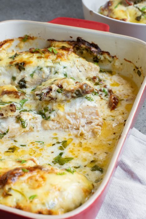 Baked Tuscan chicken casserole  in a red dish topped with melted cheese, herbs, and a creamy sauce.