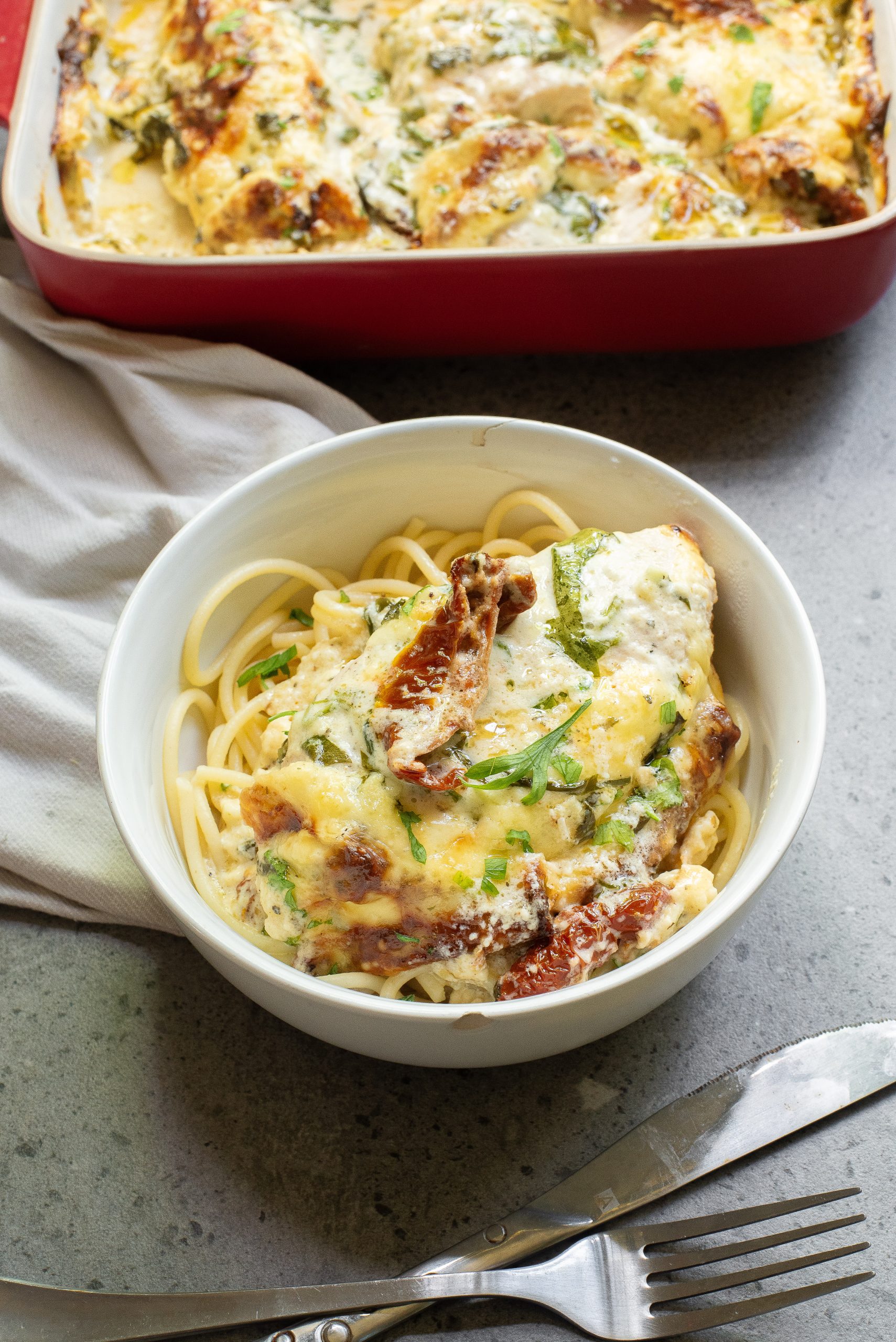 A bowl of pasta topped with a creamy, cheesy mixture of chicken, sundried tomatoes, and herbs. A similar dish in a red baking tray is in the background. Fork and knife are nearby.