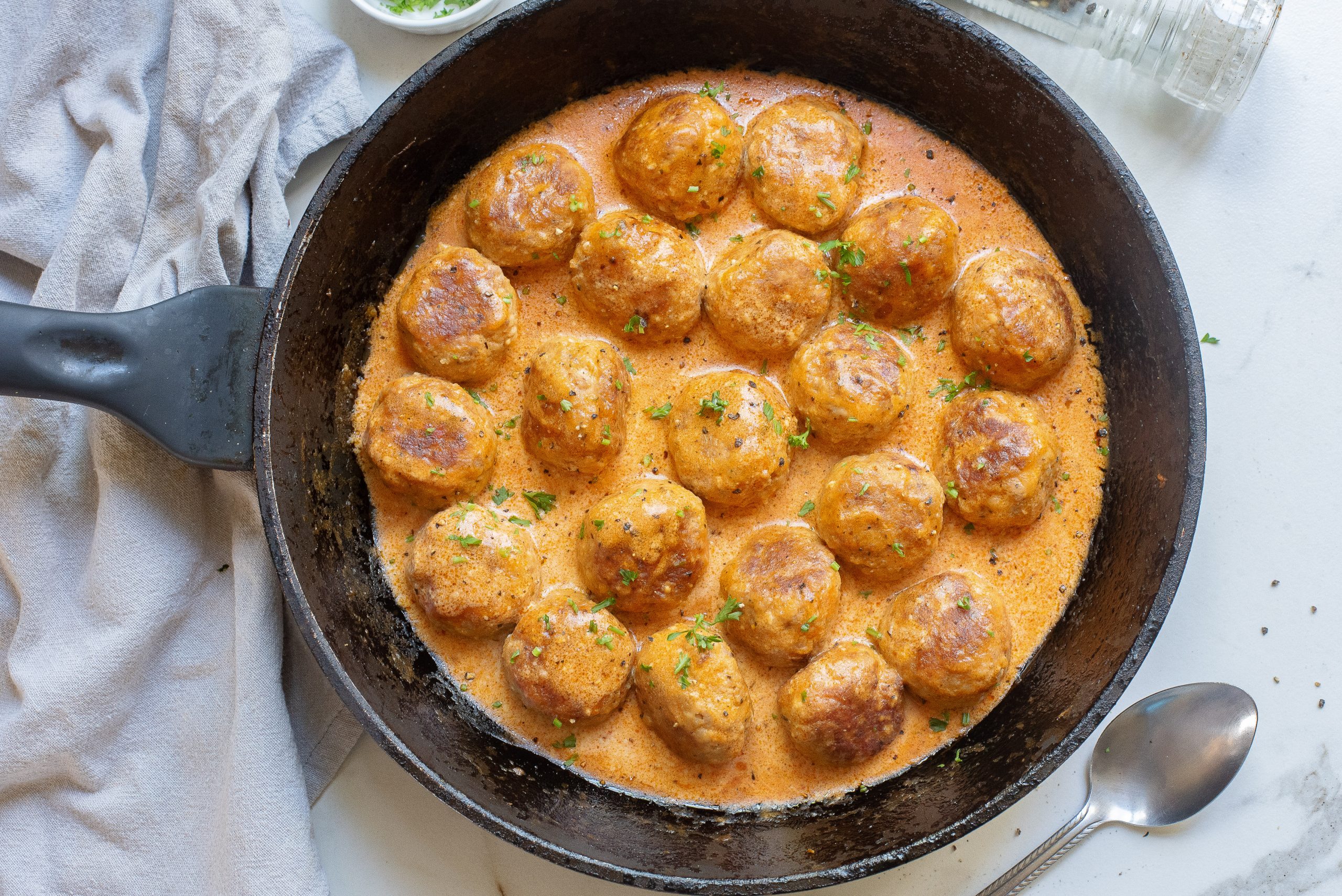 Meatballs in a creamy sauce in a black skillet, garnished with herbs. Nearby are a spoon, a light cloth, and a small bowl of herbs.