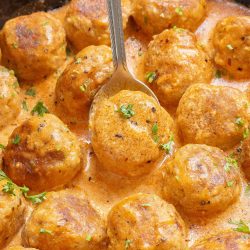 A skillet filled with cooked meatballs in a creamy sauce, garnished with chopped herbs, with a serving spoon resting among the meatballs.