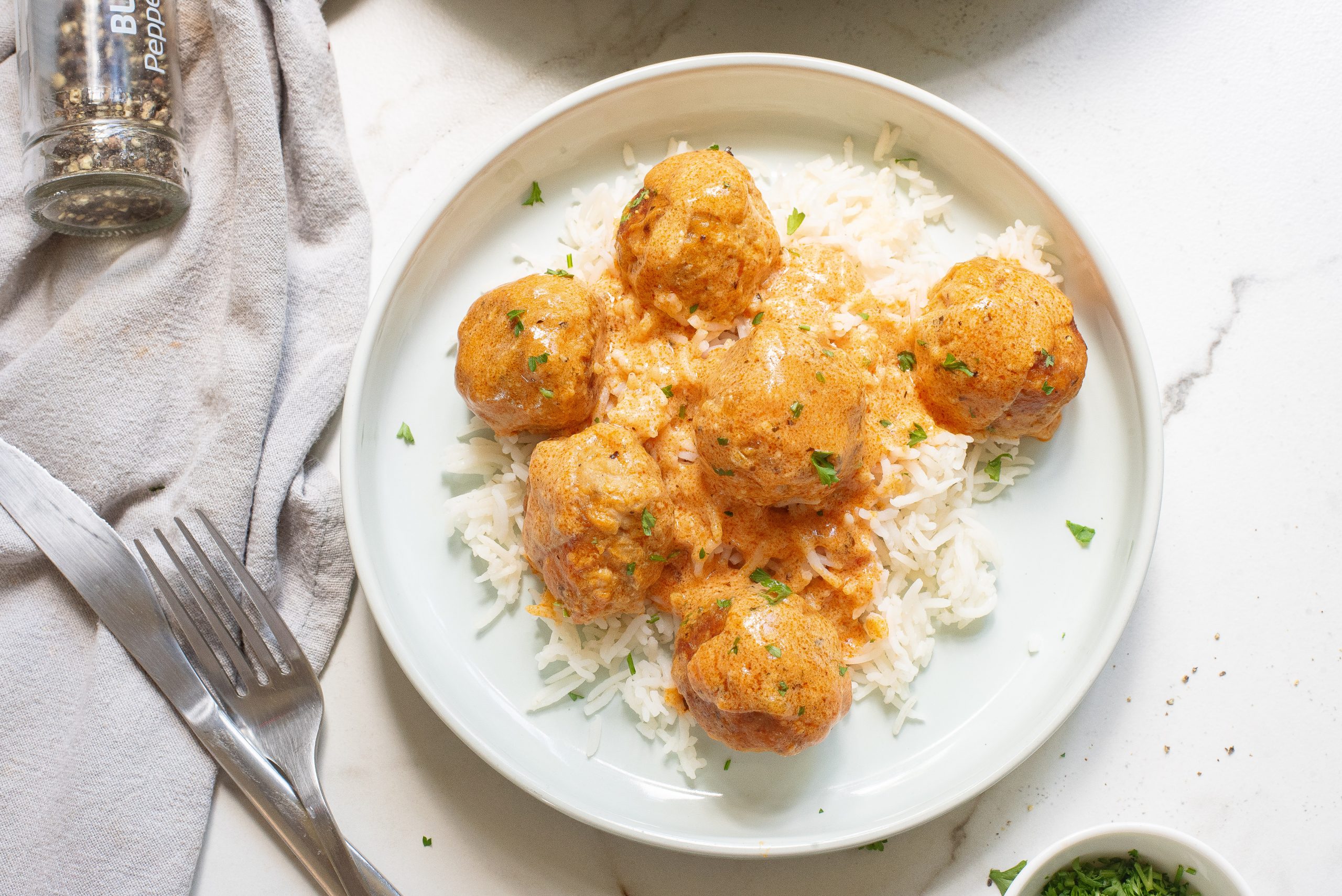 Plate of meatballs in creamy sauce served over white rice, garnished with chopped herbs, with a fork and a napkin beside the plate on a marble surface.