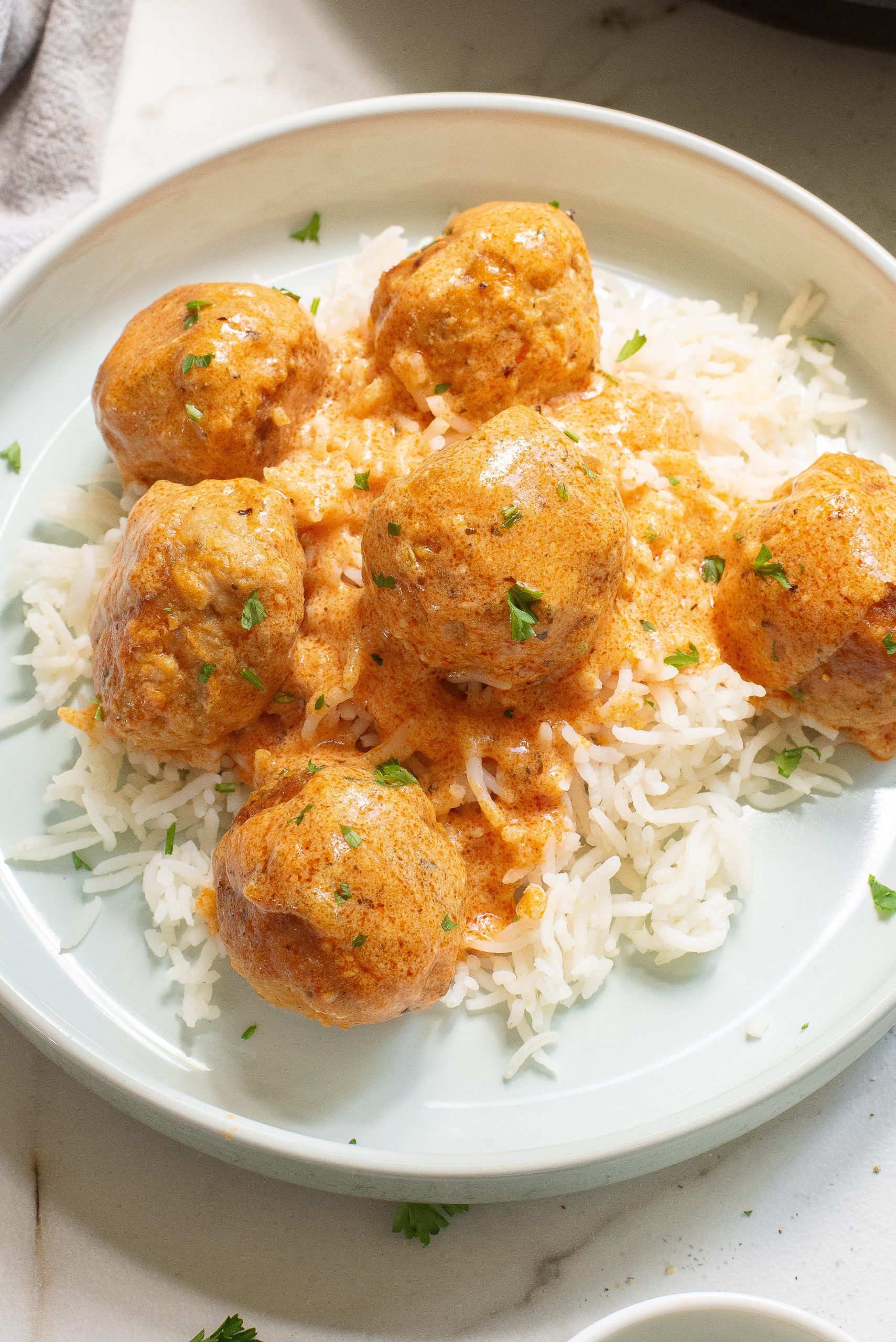 Plate of rice topped with Buffalo chicken meatballs in a creamy orange sauce, garnished with chopped herbs.