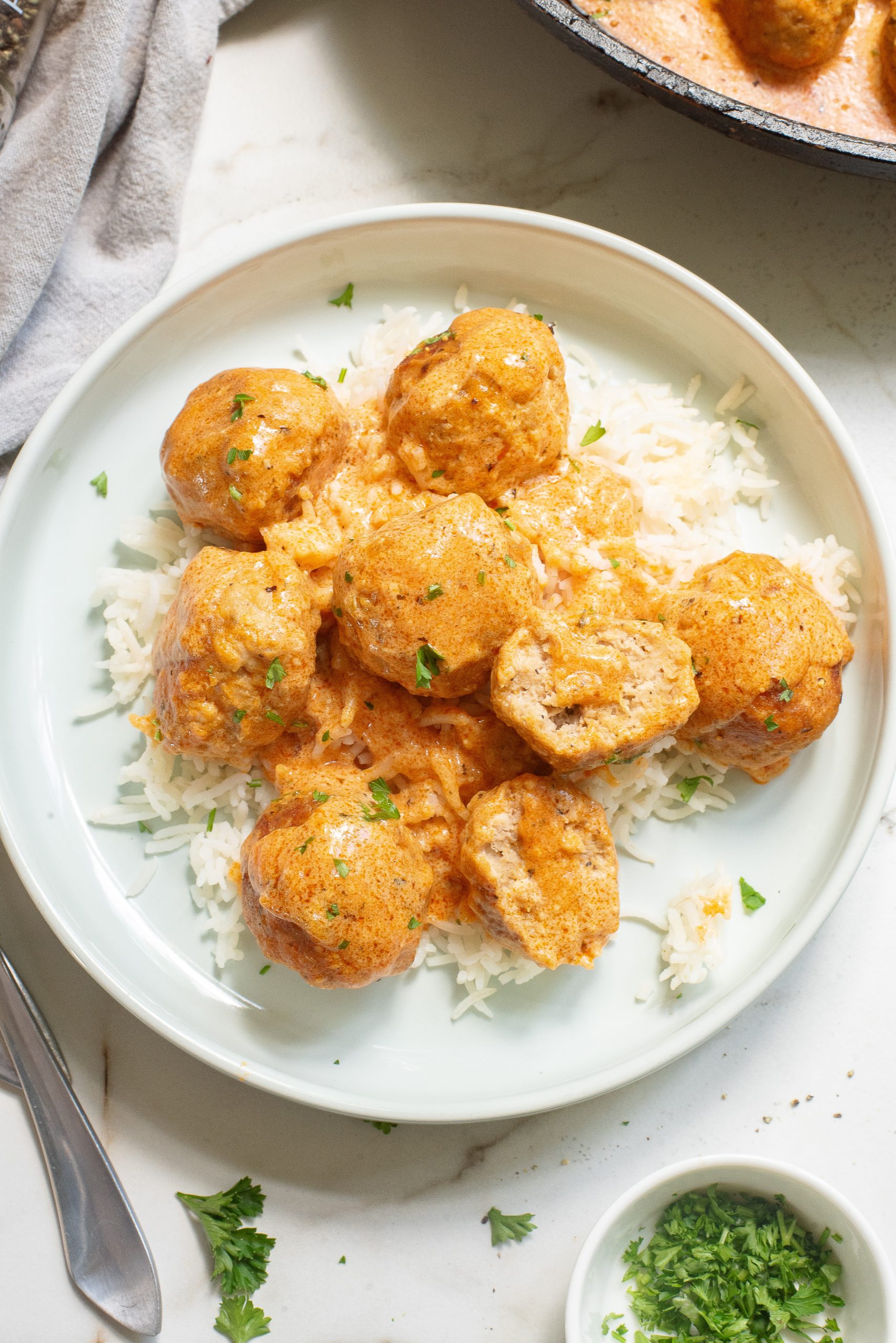 A plate of Buffalo chicken meatballs in a creamy orange sauce served over white rice, garnished with chopped herbs. A spoon and a small bowl of herbs are nearby.