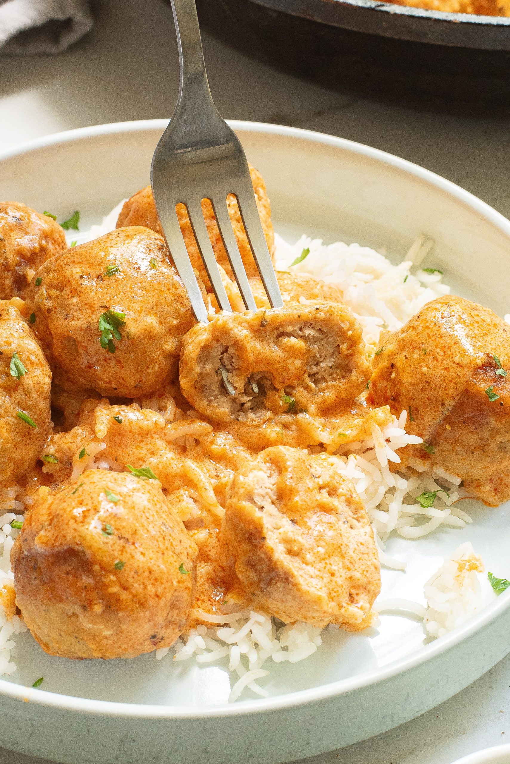 A fork cuts into a Buffalo chicken meatballs covered in creamy sauce on a plate of rice, garnished with herbs.