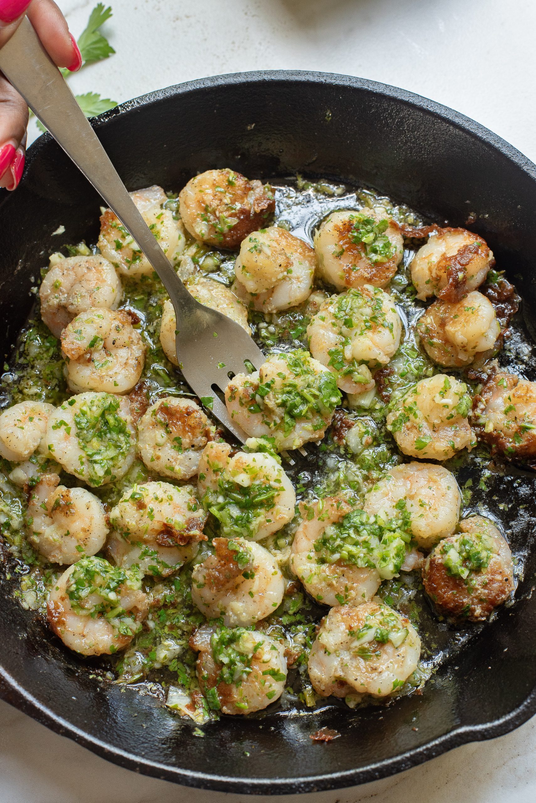 A skillet filled with cooked shrimp garnished with green herbs. A person holds a fork, poised to pick up a shrimp.