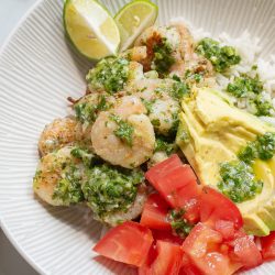 A bowl of shrimp with green sauce, rice, avocado slices, diced tomatoes, and lime wedges.