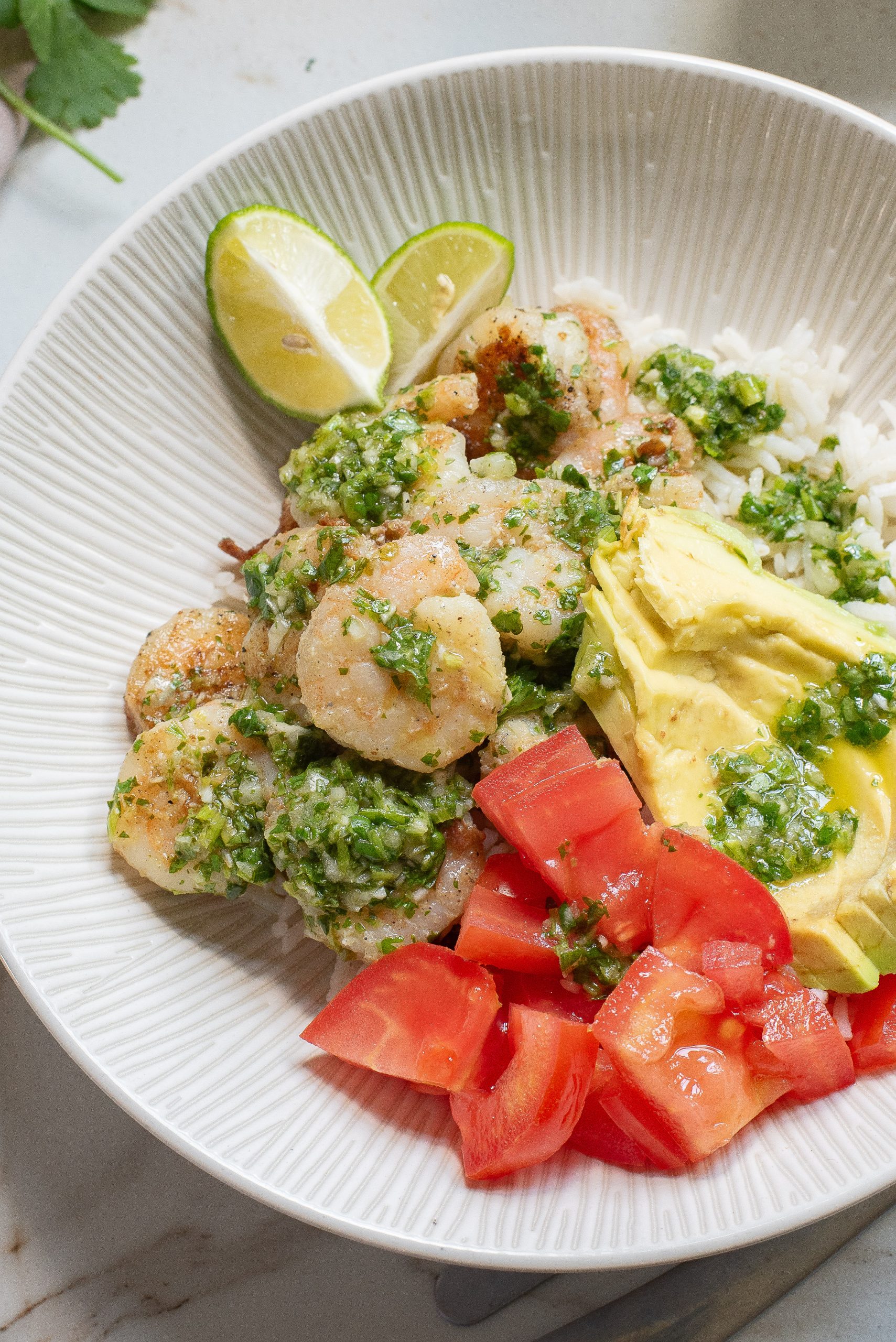 A bowl of shrimp with green sauce, rice, avocado slices, diced tomatoes, and lime wedges.