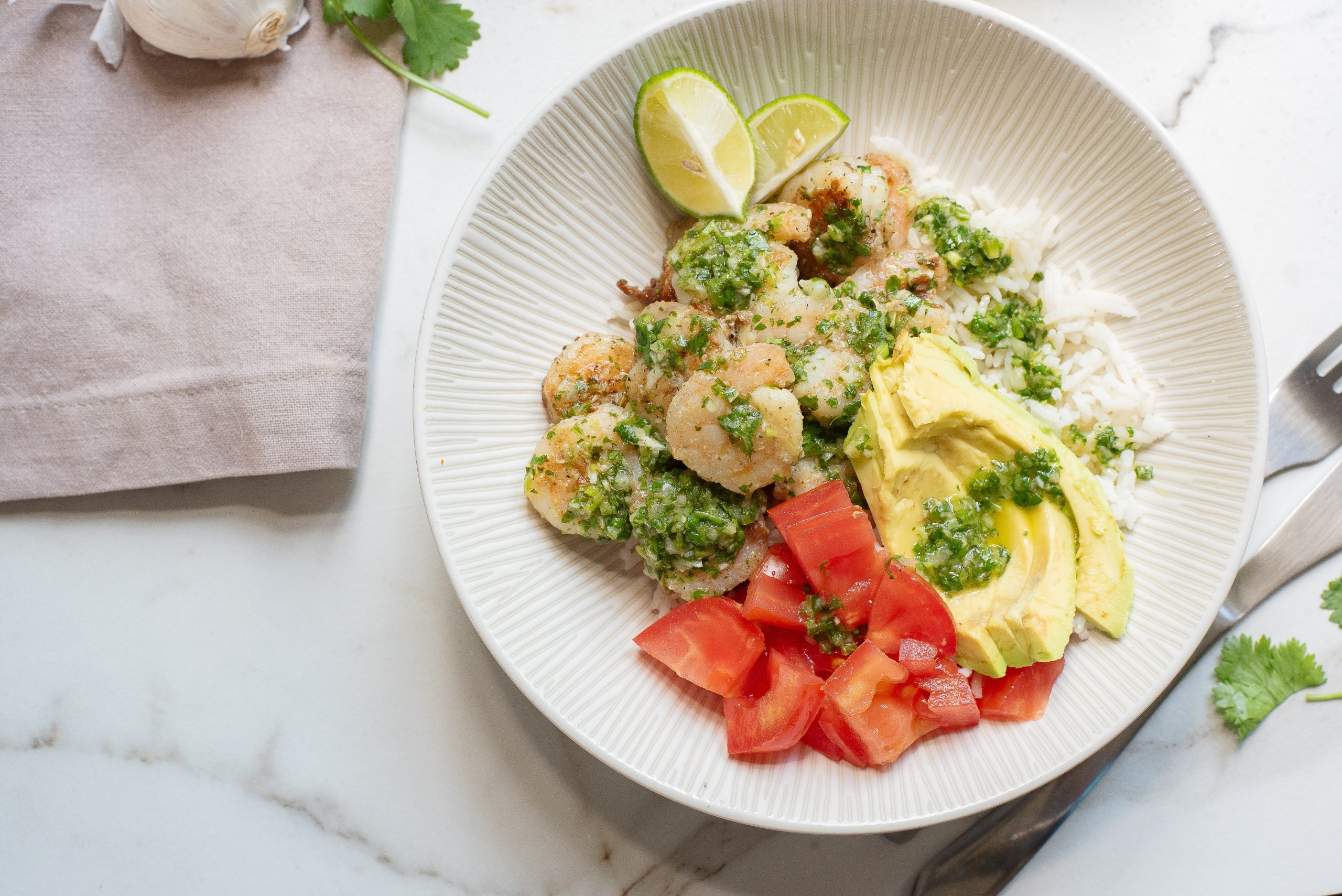 A plate with shrimp, avocado, chopped tomatoes, white rice, and chimichurri sauce, garnished with lime wedges.