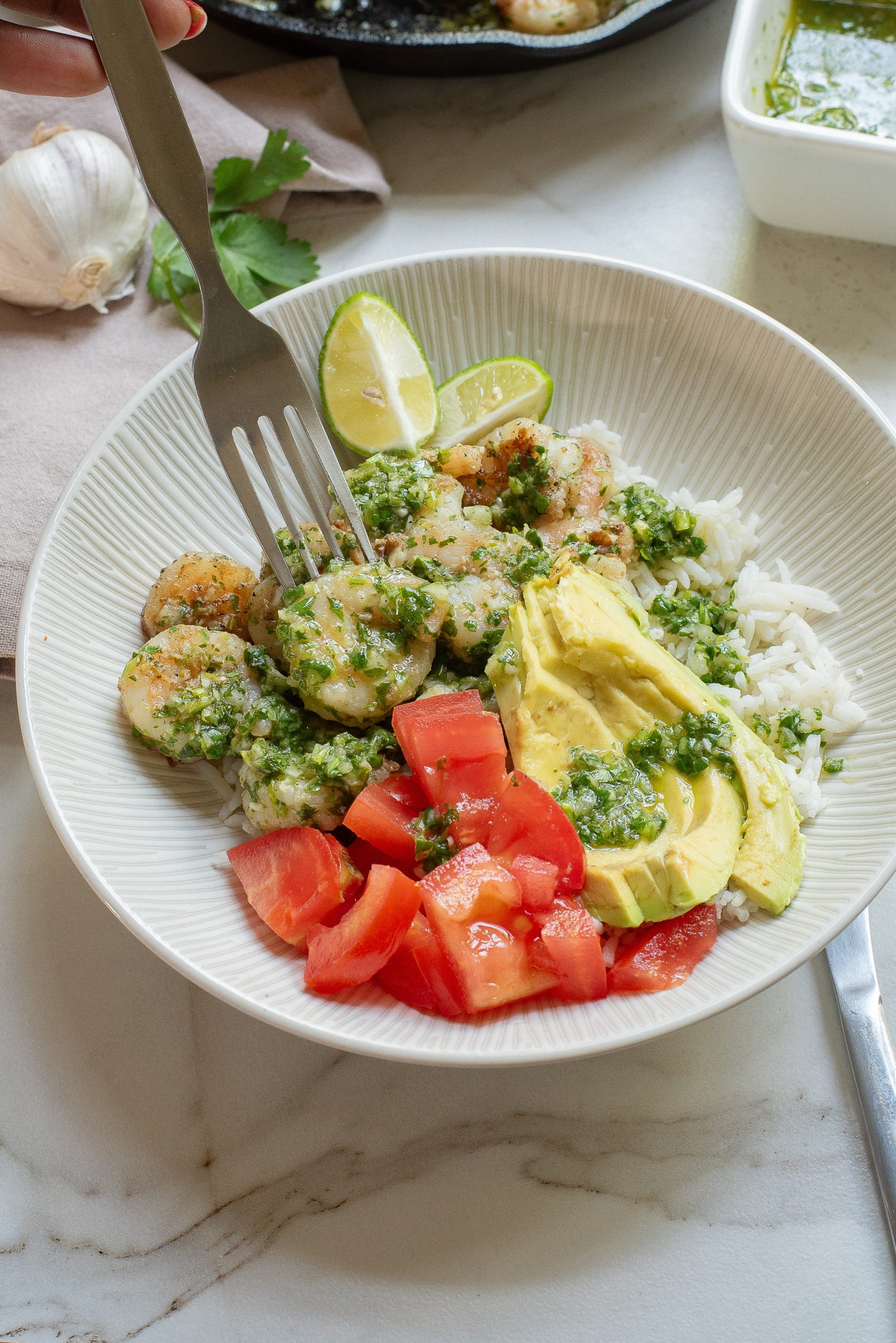 A bowl of rice topped with shrimp, avocado slices, diced tomatoes, and green sauce, garnished with lime wedges. A fork is placed in the bowl, and garlic and sauce are nearby.