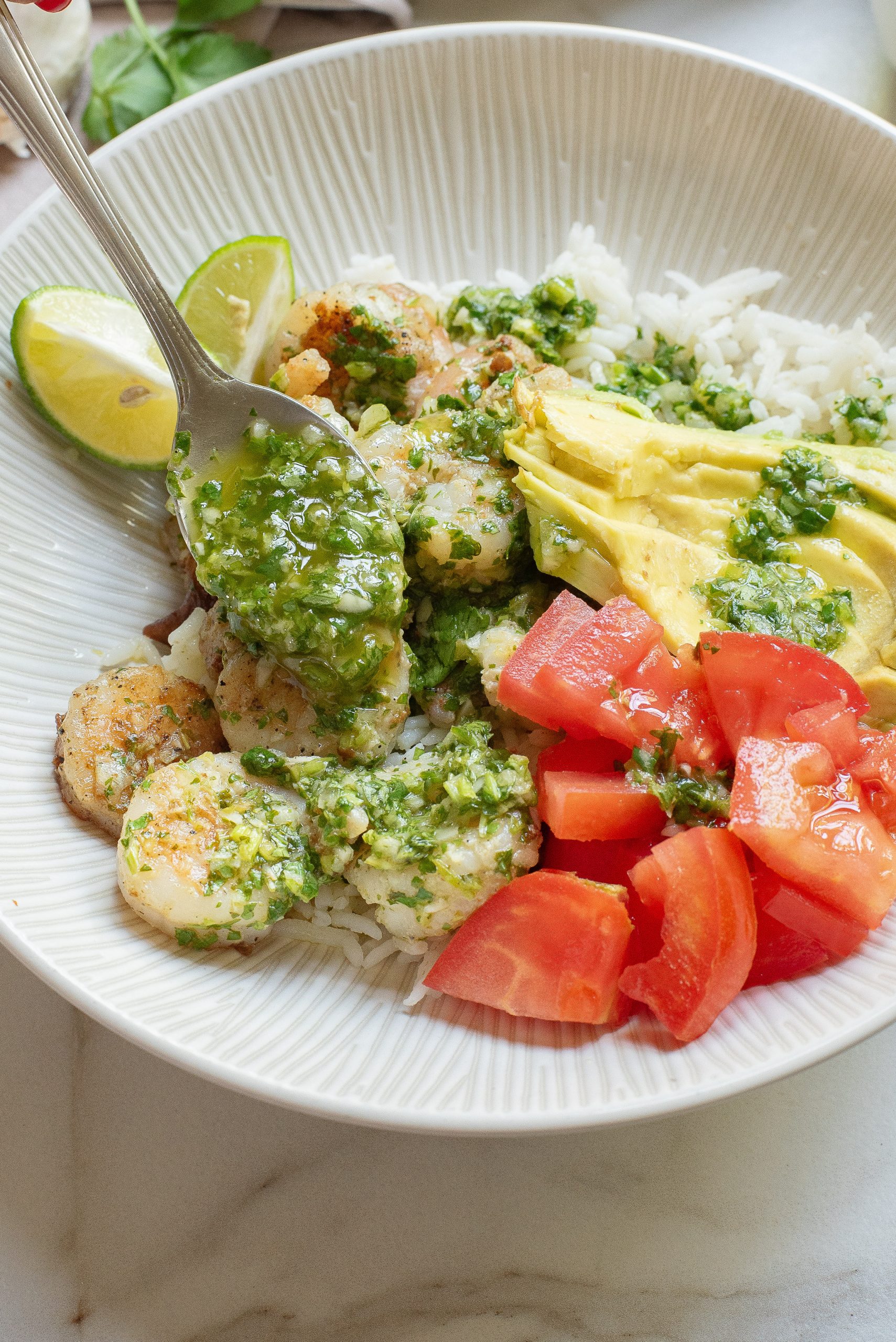 A bowl with rice, shrimp, avocado slices, diced tomatoes, lime wedges, and green sauce on top.