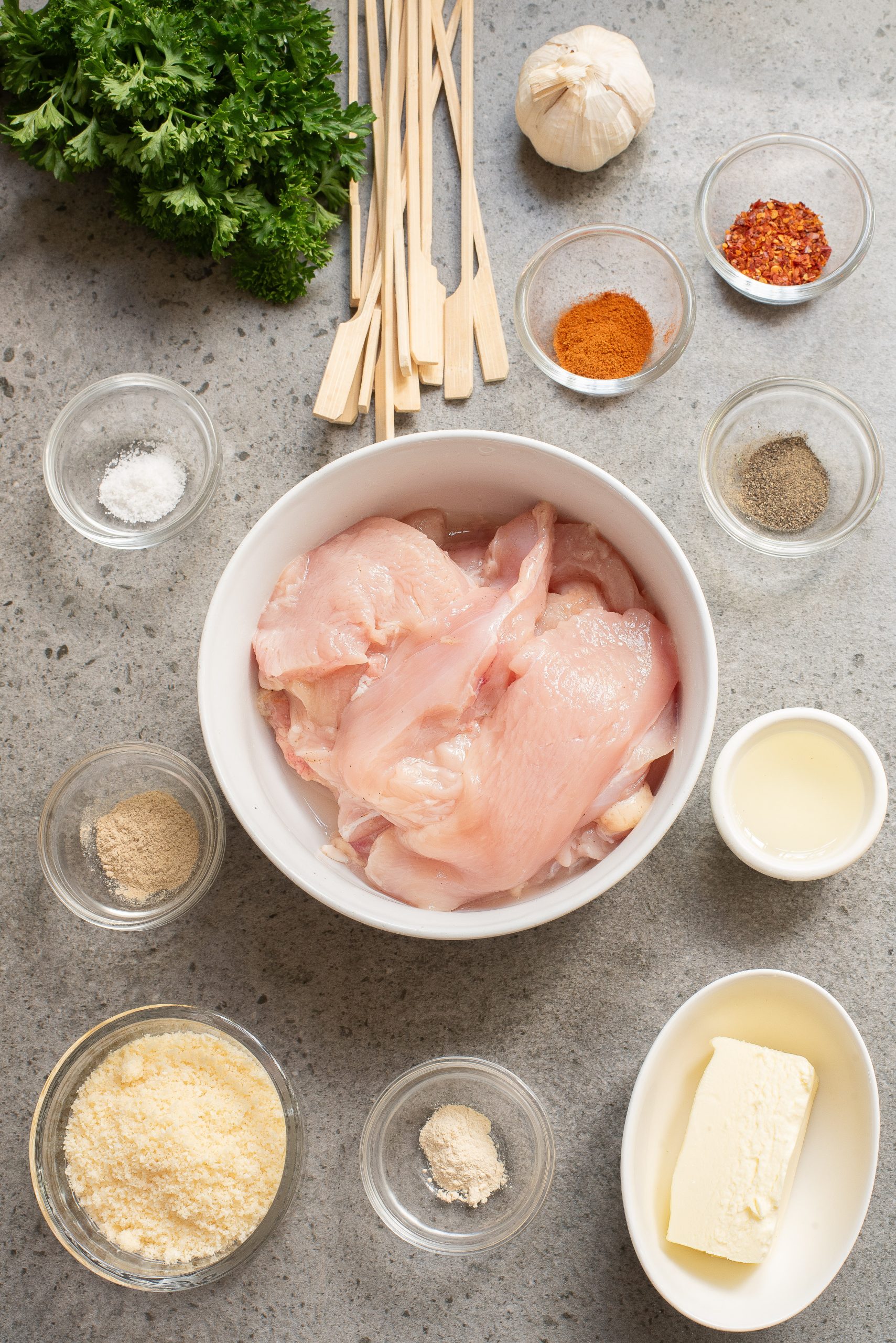 Raw chicken breast pieces in a bowl surrounded by ingredients: butter, garlic, seasonings, skewers, and parsley on a gray surface.