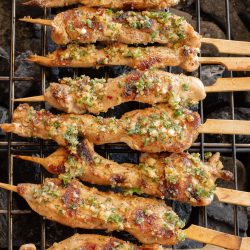 Skewers of marinated chicken grilling over charcoal, topped with chopped herbs and garlic.