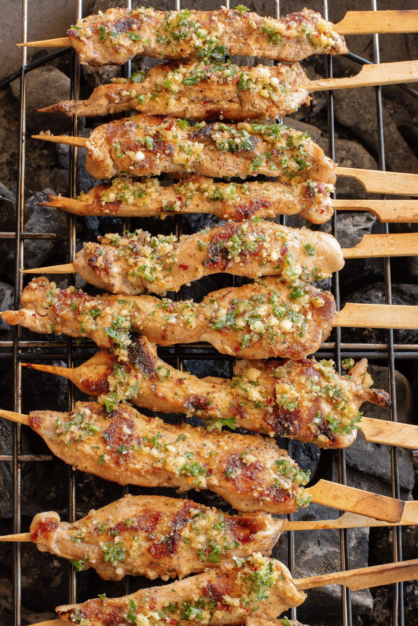 Skewers of marinated chicken grilling over charcoal, topped with chopped herbs and garlic.