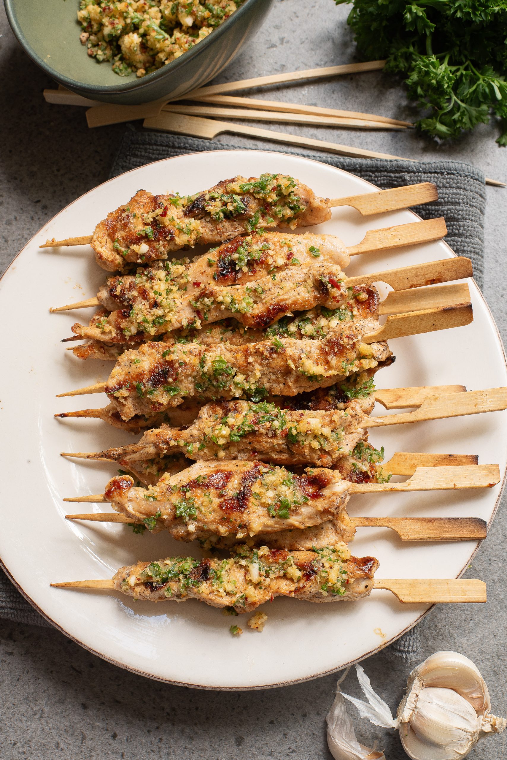 Grilled chicken skewers topped with a textured herb and spice mixture on a white plate. Garlic and fresh herbs are beside the plate.