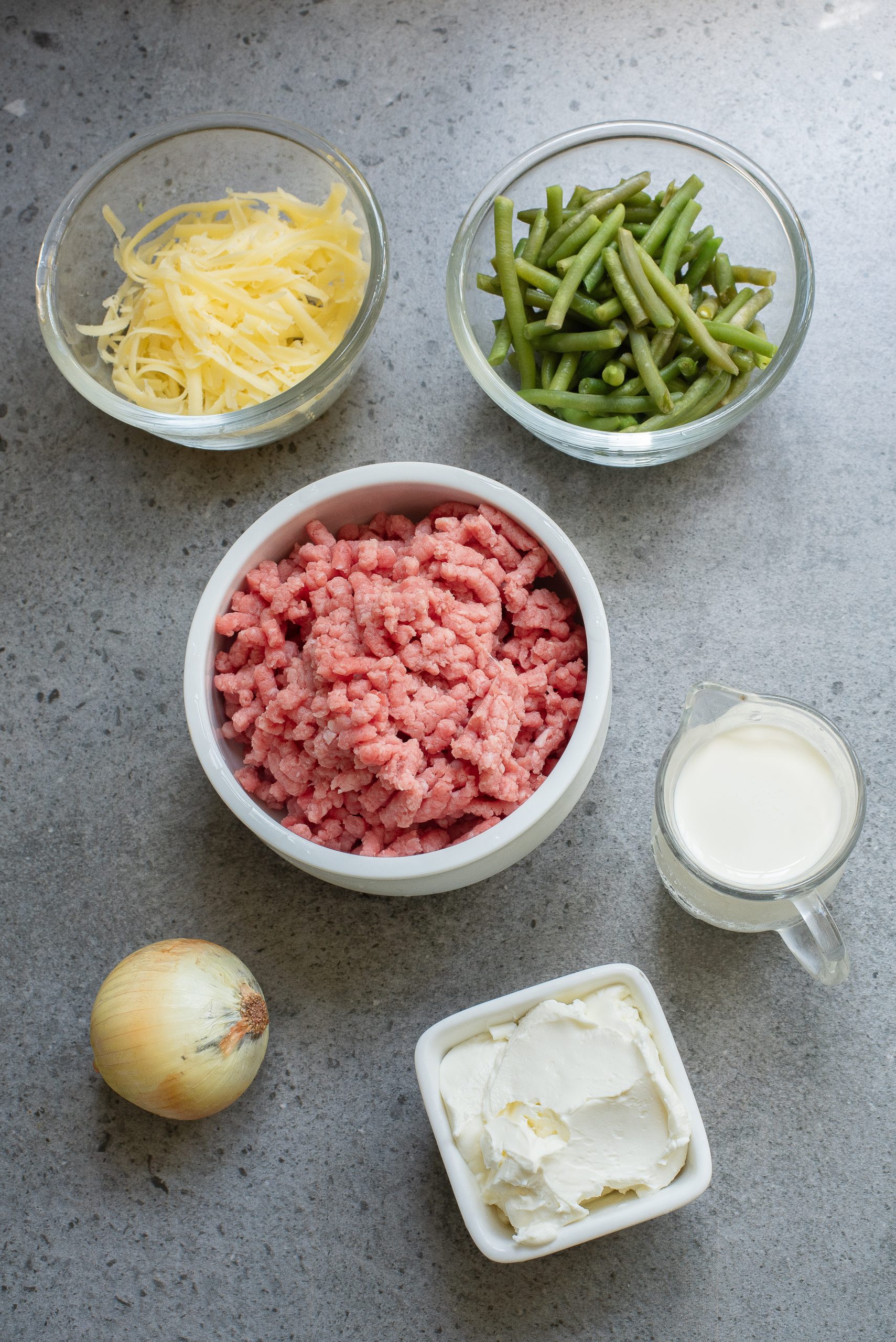 Ground meat, shredded cheese, green beans, milk, cream cheese, and an onion arranged on a gray countertop.