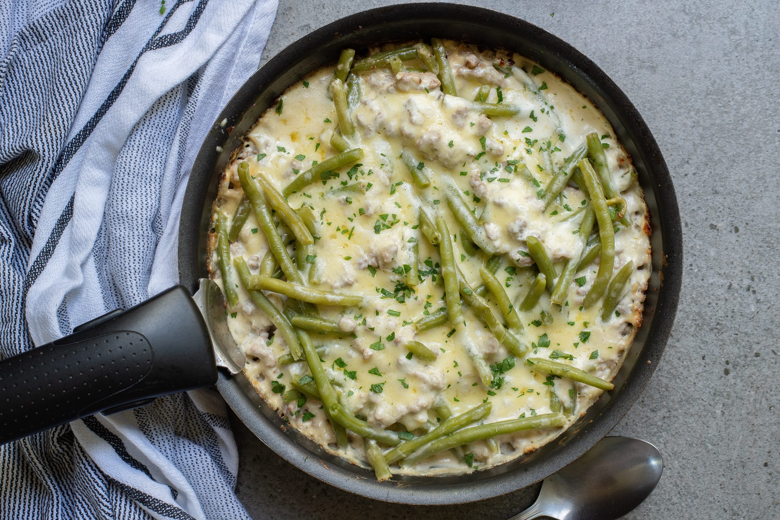 A Lazy Keto Ground Beef Recipe with creamy green bean casserole topped with melted cheese and chopped herbs. A striped cloth is nearby.