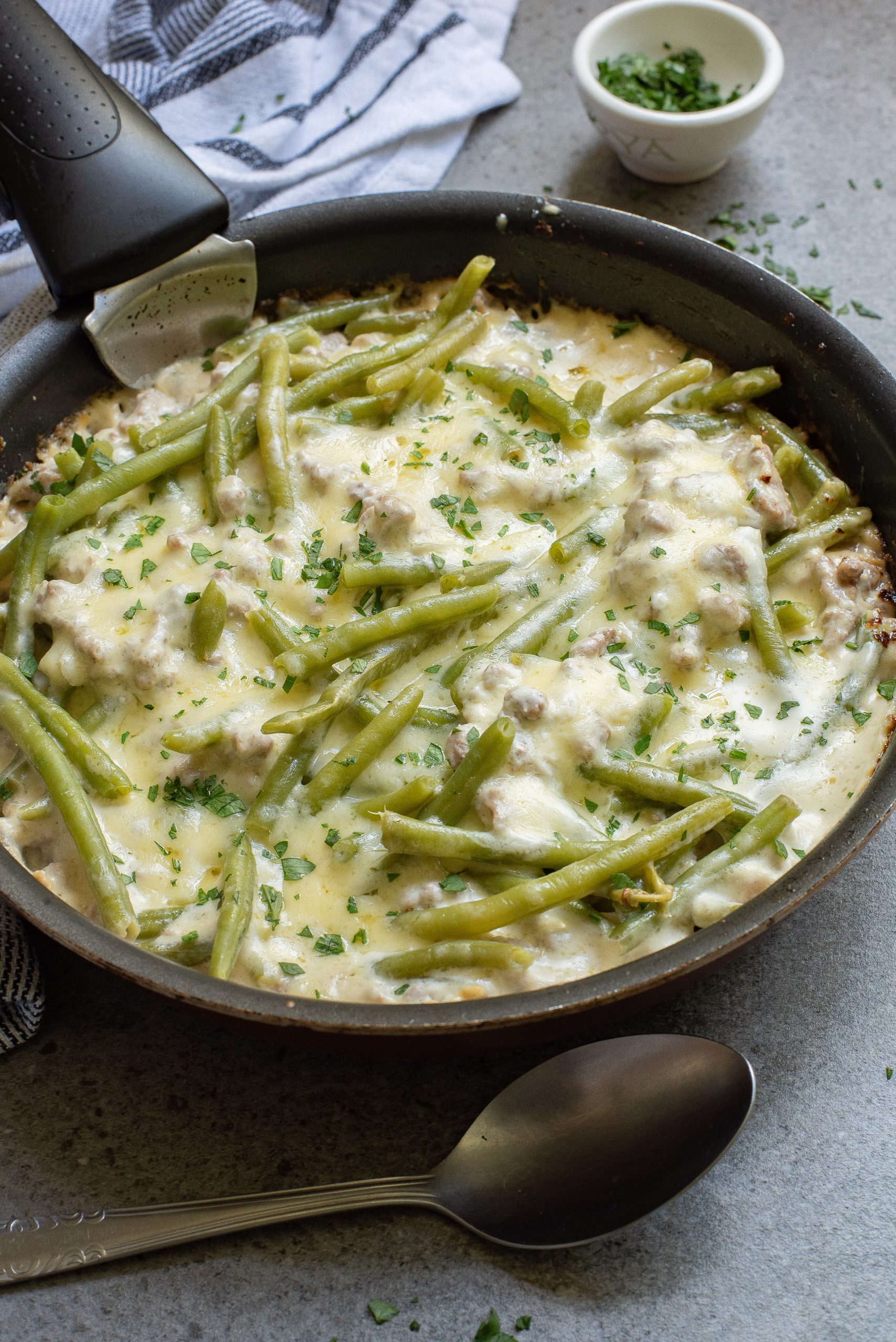 A pan of green bean casserole topped with melted cheese and herbs, with a spoon beside it on a gray surface. A small bowl of chopped herbs is in the background.