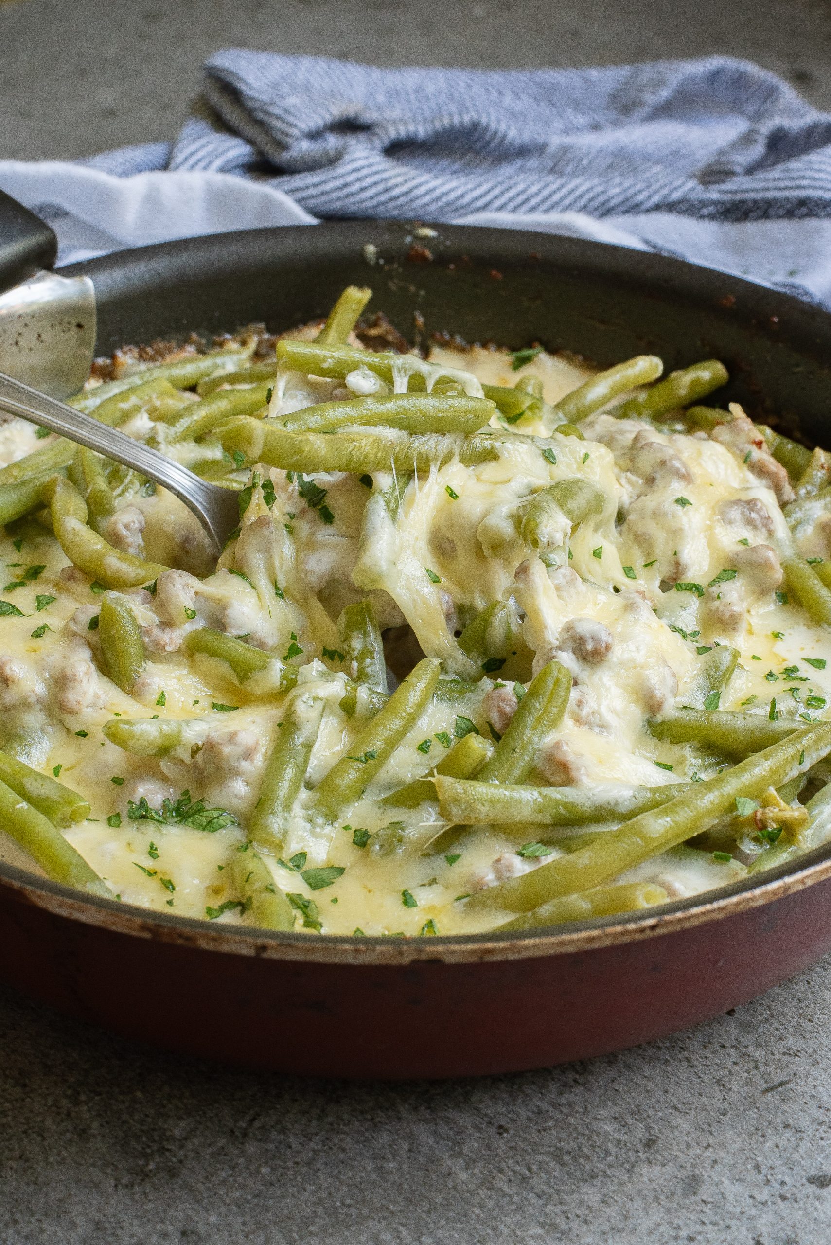 A Lazy Keto Ground Beef Recipe with cooked green beans and melted cheese, garnished with herbs, and a serving spoon resting inside.