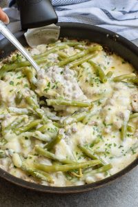 A skillet with green beans, ground meat, and melted cheese, garnished with herbs, being stirred with a spoon.