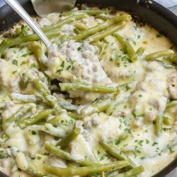 A skillet with green beans, ground meat, and melted cheese, garnished with herbs, being stirred with a spoon.