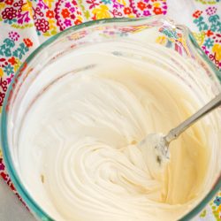 A glass bowl filled with smooth, creamy mixture, with a metal spoon, on a colorful, patterned cloth.