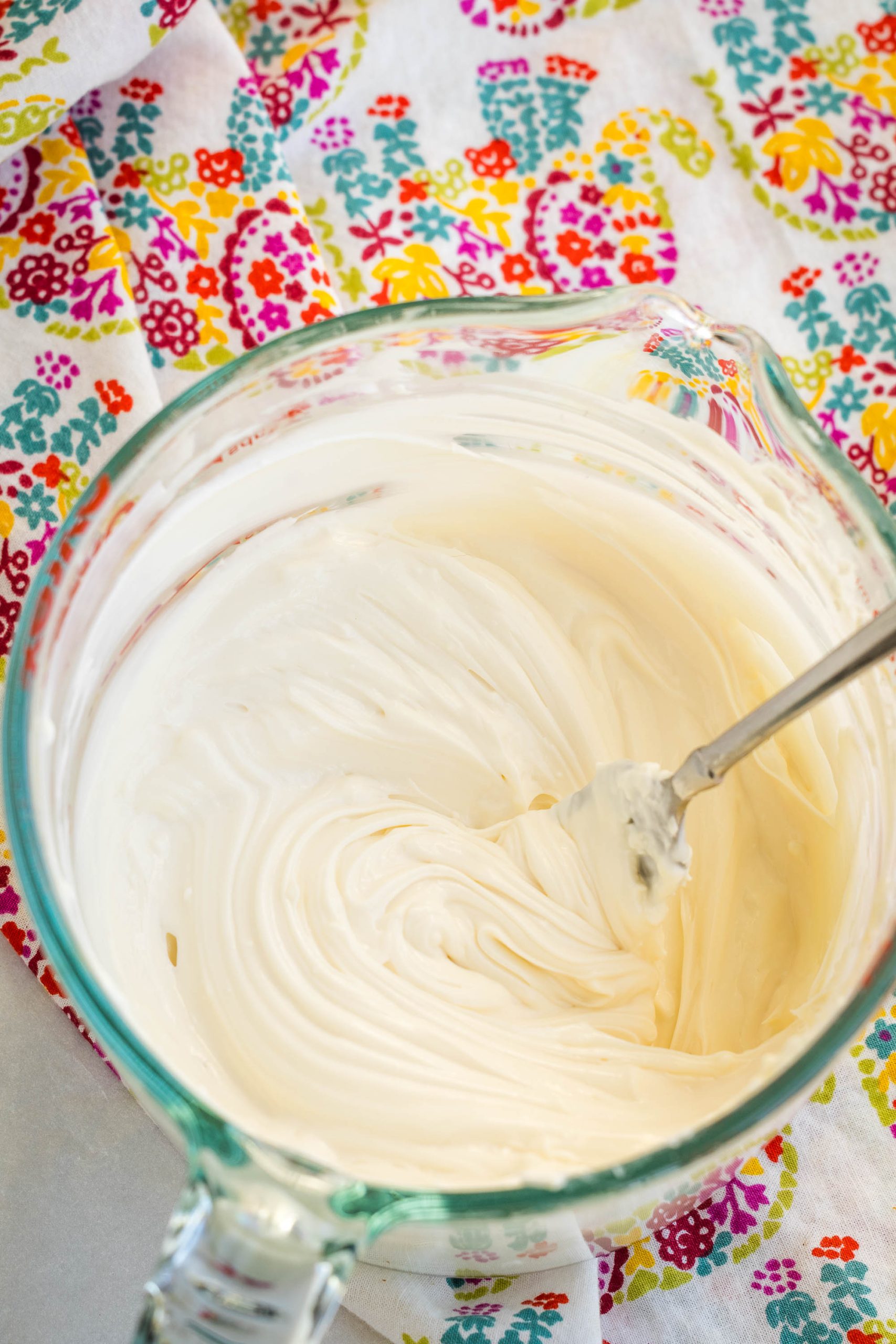 A glass bowl filled with smooth, creamy Healthy Cream Cheese Frosting, with a metal spoon, on a colorful, patterned cloth.