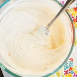 A glass bowl with smooth, creamy frosting being mixed by a metal spoon, placed on a colorful floral-patterned cloth.