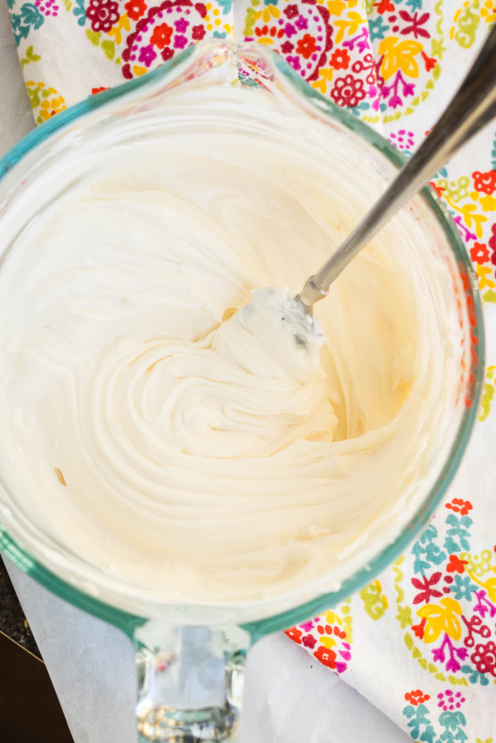 A glass bowl with smooth, creamy frosting being mixed by a metal spoon, placed on a colorful floral-patterned cloth.