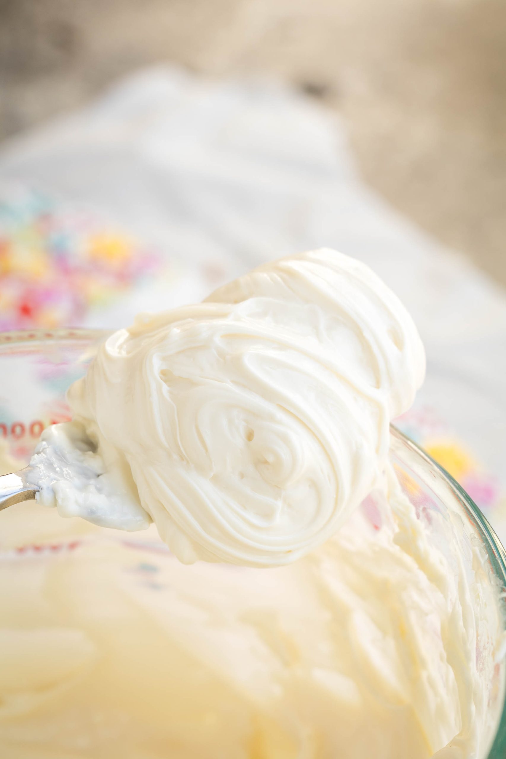 A spoon holds a dollop of thick, smooth white Healthy Cream Cheese Frosting above a glass bowl filled with more of the same creamy substance. The background is softly blurred.